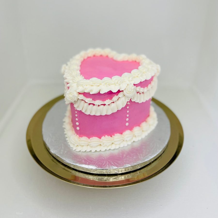 A vibrant pink heart-shaped cake featuring intricate white ruffled buttercream piping along the edges and small white pearl details on the s