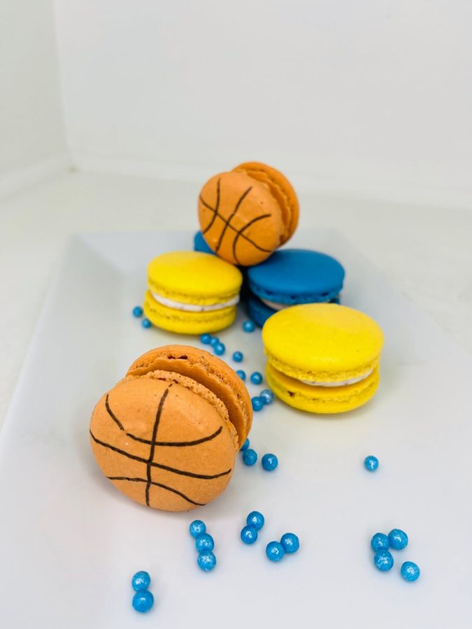 Custom basketball themed orange macarons, alongside yellow and blue macarons, decorated with blue sprinkles on a white platter.