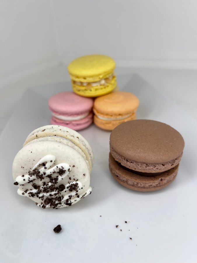 A vibrant assortment of macarons featuring yellow, pink, orange, white, and chocolate flavors, with one white macaron decorated with white d