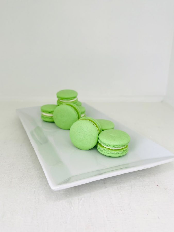 A collection of light green macarons with white filling is neatly arranged on a white rectangular serving plate, presented against a clean w