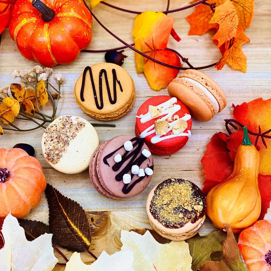 A festive assortment of fall-themed macarons in various colors and flavors, decorated with chocolate drizzles, marshmallows, and crushed top