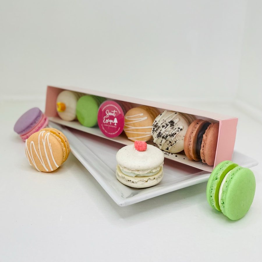 A vibrant assortment of macarons featuring various colors and decorations, including white drizzle, cookie crumbs, a branded logo, and a sma
