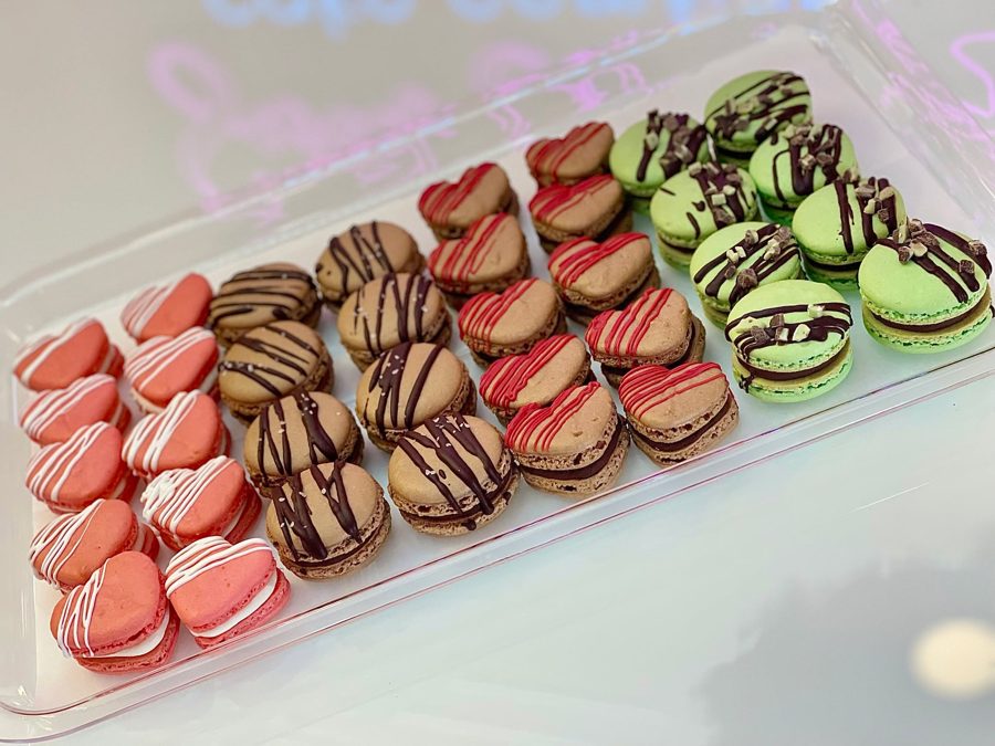 A clear tray filled with an assortment of heart-shaped and round macarons in pink, green, and brown, decorated with white, red, and chocolat