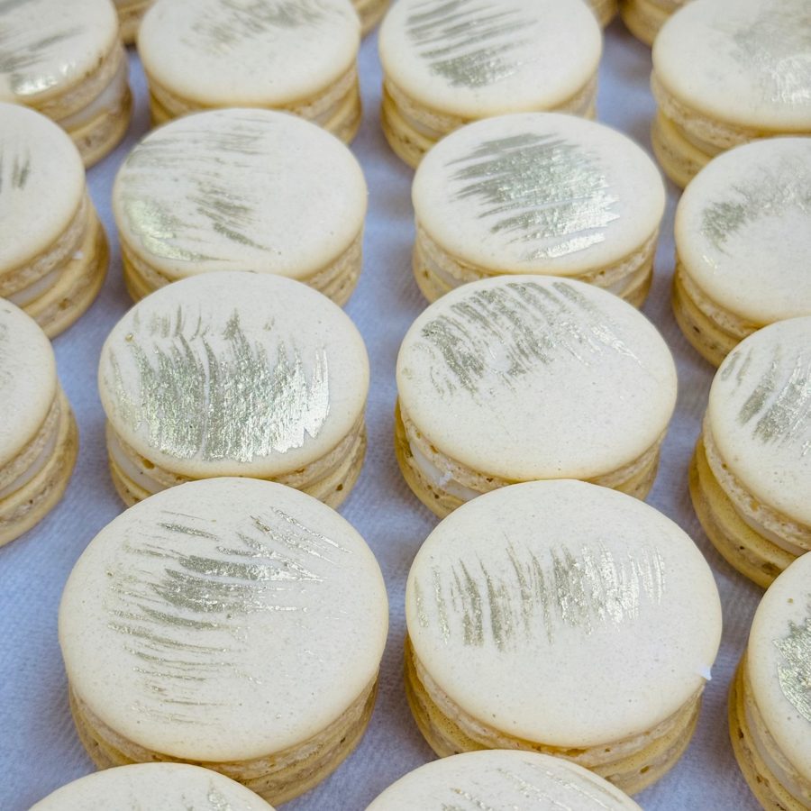 Elegant cream-colored macarons adorned with delicate gold brushstroke decorations, ideal for sophisticated events and dessert bars.