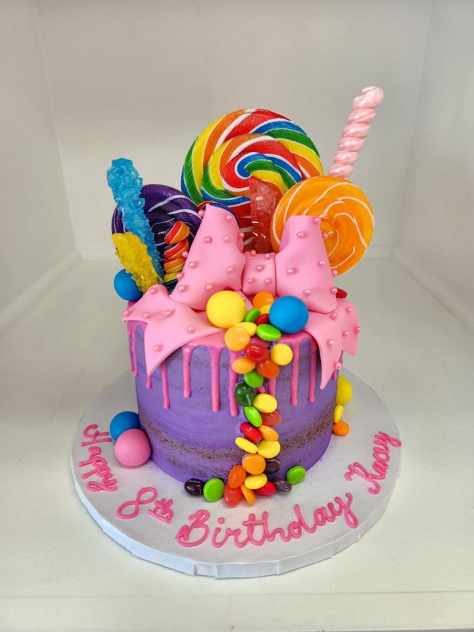 A vibrant single-tier purple drip cake adorned with a pink fondant bow, various lollipops, rock candy, and colorful candies, featuring "Happ